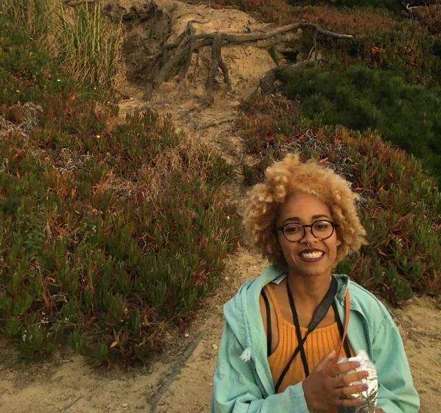 Akaelah looks at the camera, smiling, holding a small package in her hands. They are wearing glasses, dark red lipstick, and their blonde hair is very curly and thick. They are wearing a yellow tank top and aqua jacket. Behind them is a sand dune covered in ice plants.